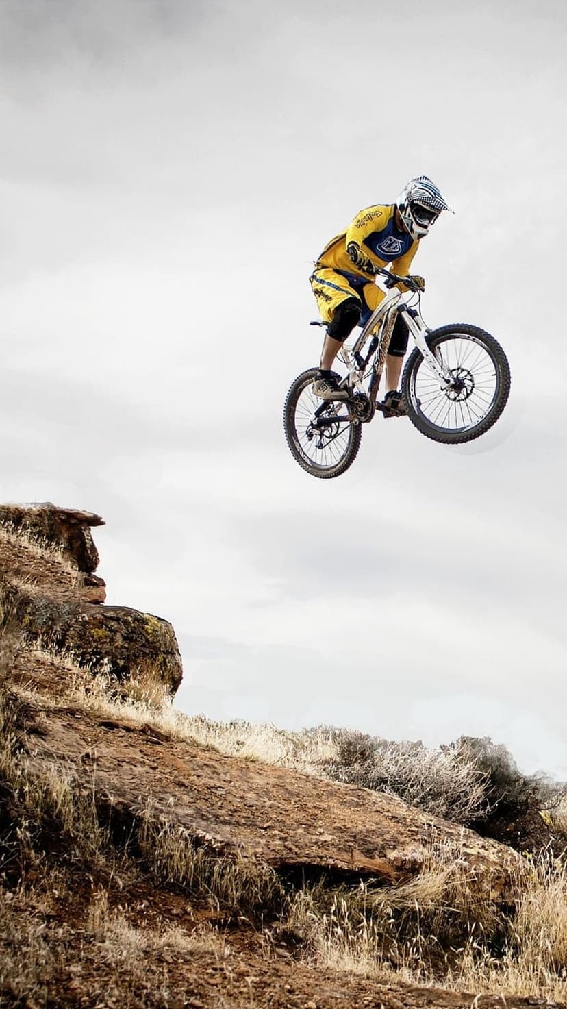 Mountain biker in a yellow jersey jumps a bicycle over a rocky ledge against a cloudy gray sky backdrop.