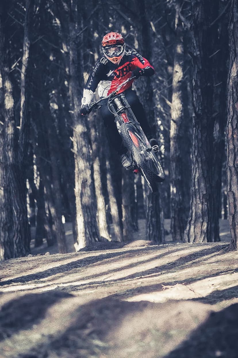 Mountain biker in mid-air jump wearing a helmet and goggles within a dark, sun-streaked coniferous forest trail.