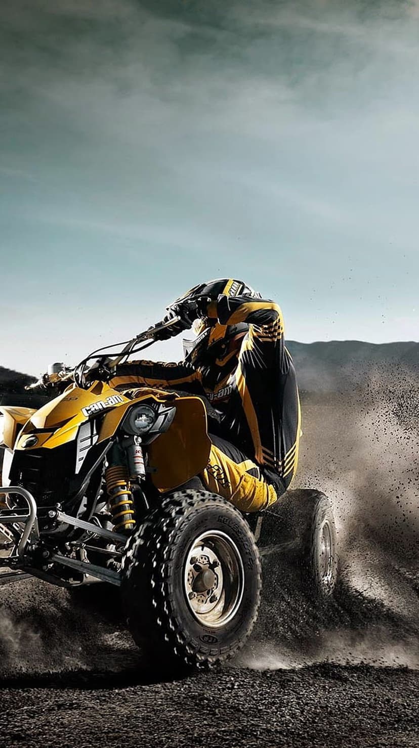 Rider in yellow gear leans a yellow quad bike into a sharp dirt turn, kicking up dust under an overcast sky.