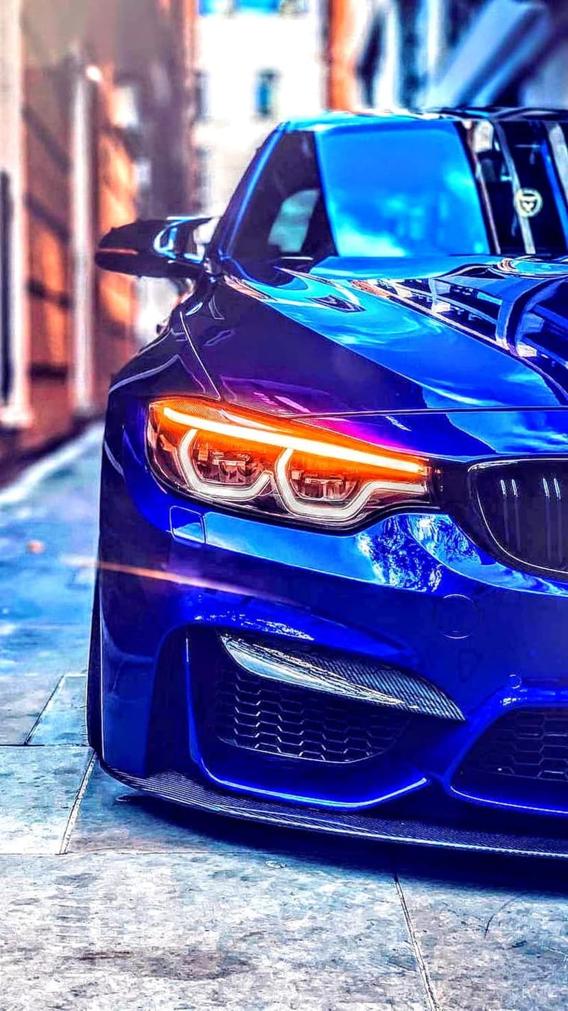 BMW M4 front headlights with glowing orange accents on a blue sports car parked on textured urban asphalt.