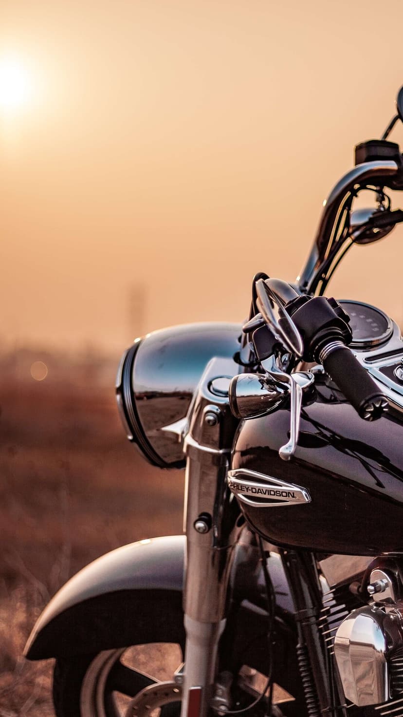 Close-up of a Harley-Davidson motorcycle headlight and chrome handlebars against a soft golden sunset glow.