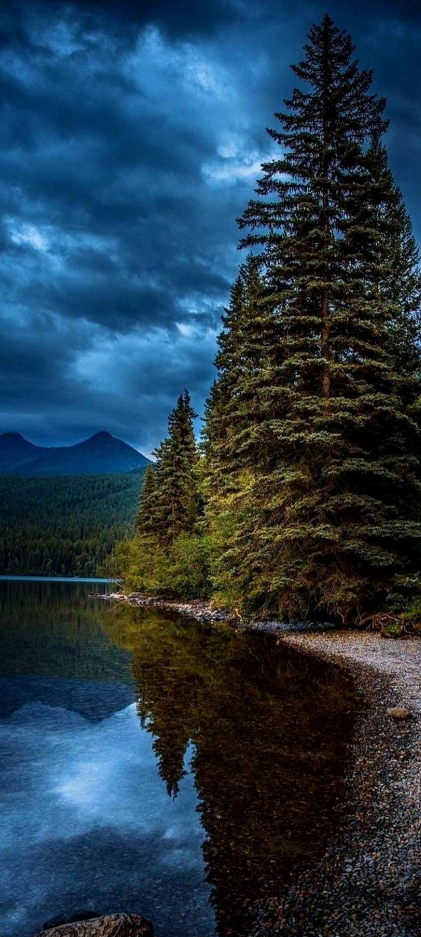 Dark storm clouds hanging over a glass-like lake reflecting a dense pine forest and distant mountain range.