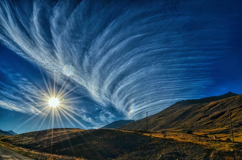 Cirrus clouds swirl in a wide arc above dark mountain silhouettes as a bright sunburst shines from the horizon.