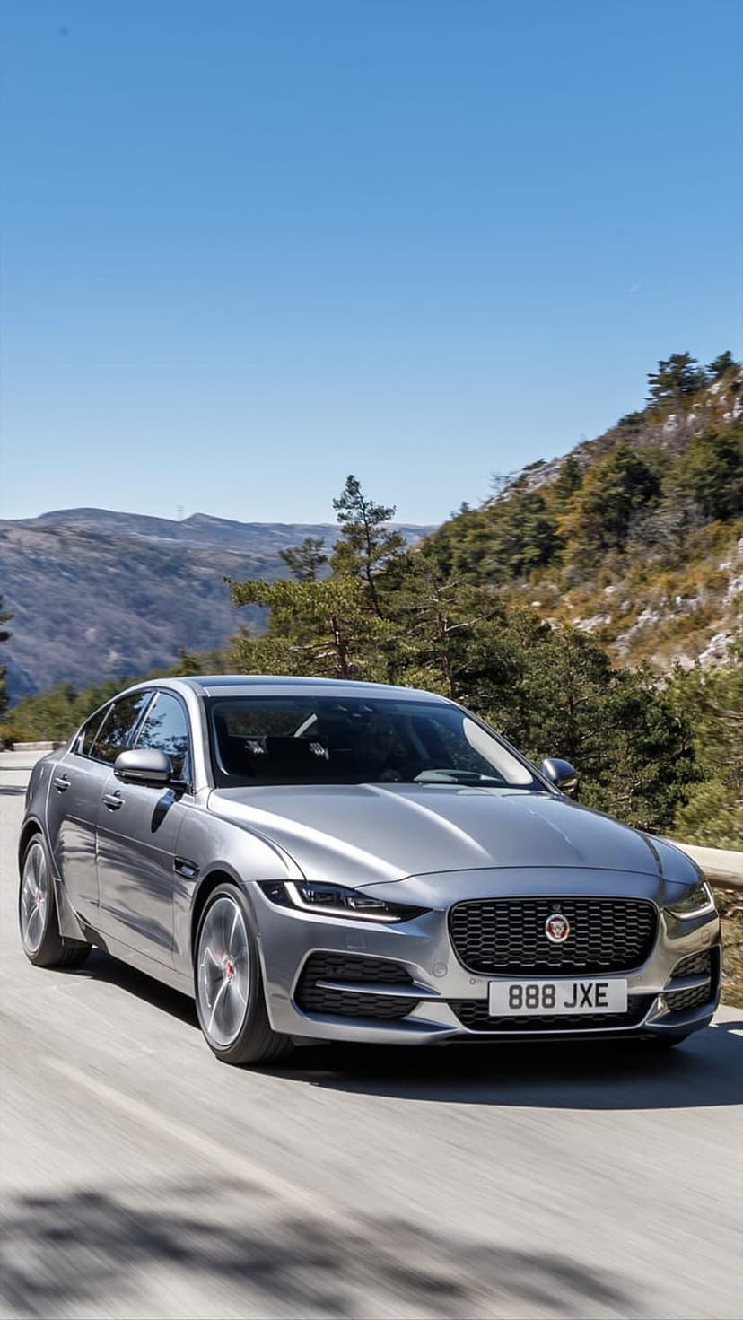 Silver Jaguar XE sedan driving on a curving mountain road surrounded by pine trees under a clear blue sky.