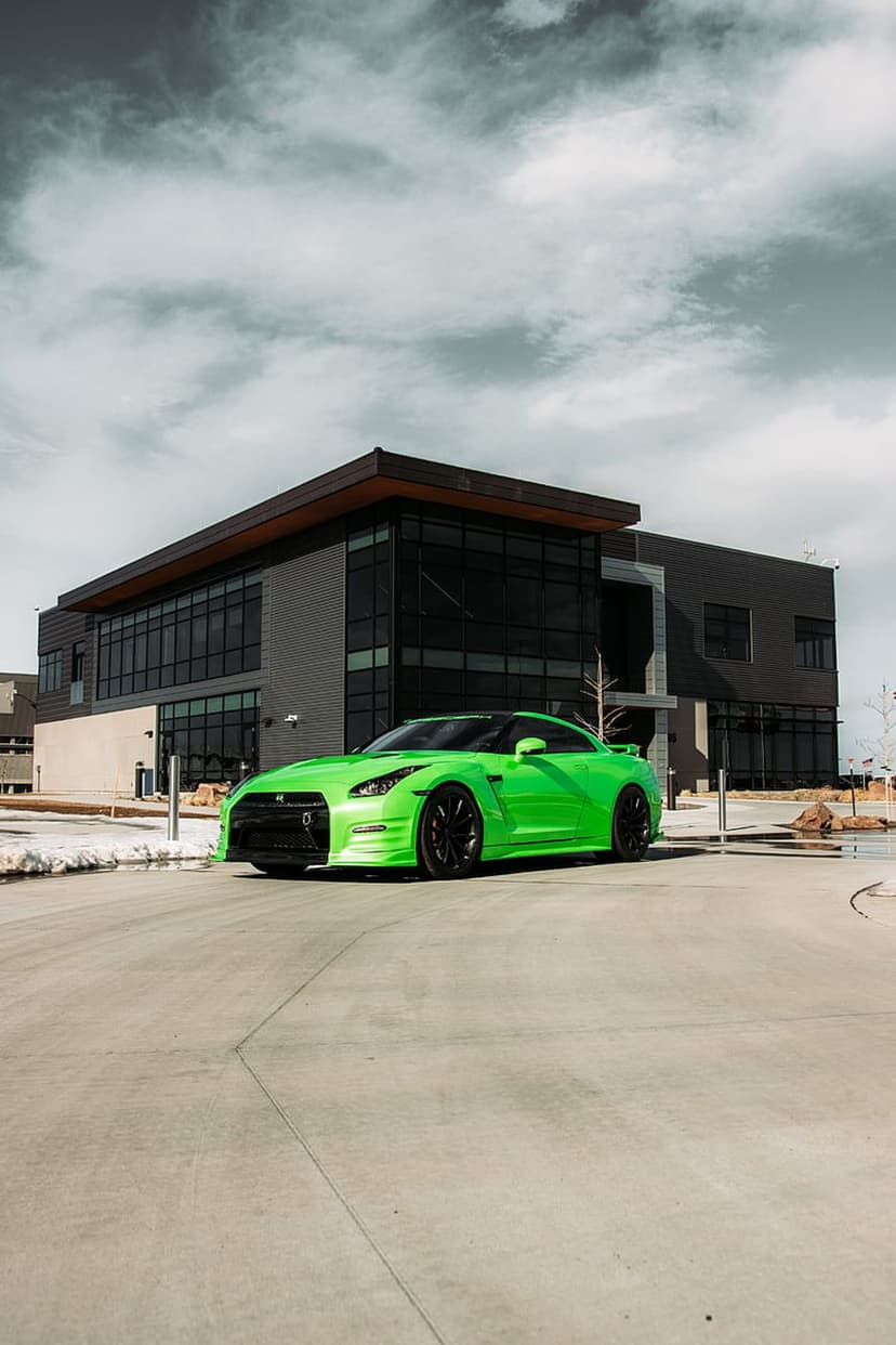 Lime green Nissan GT-R sports car parked on a concrete driveway in front of a modern glass building under grey clouds.