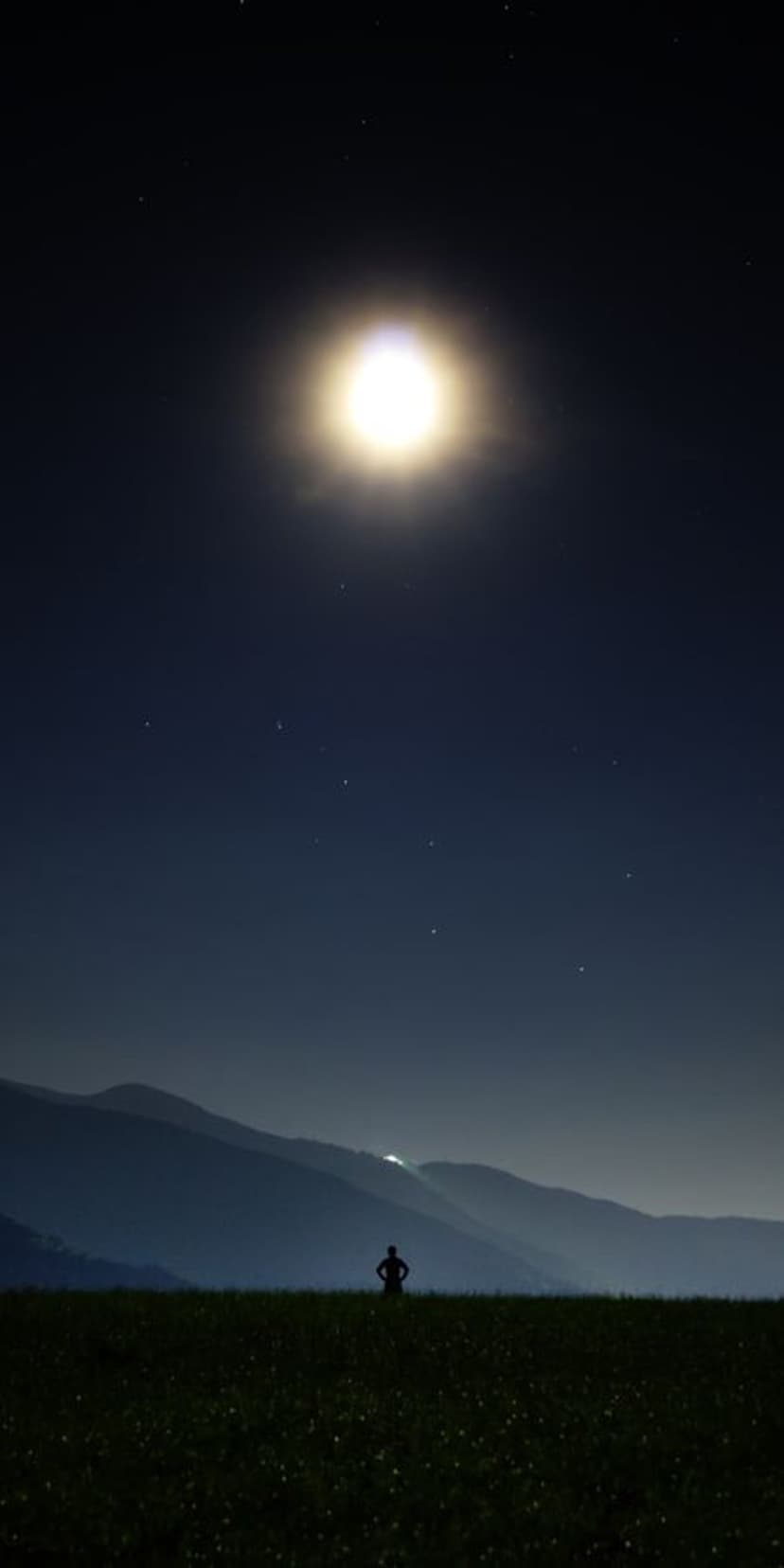 Lone person silhouette standing in a dark field beneath a massive glowing full moon and a starry night sky.