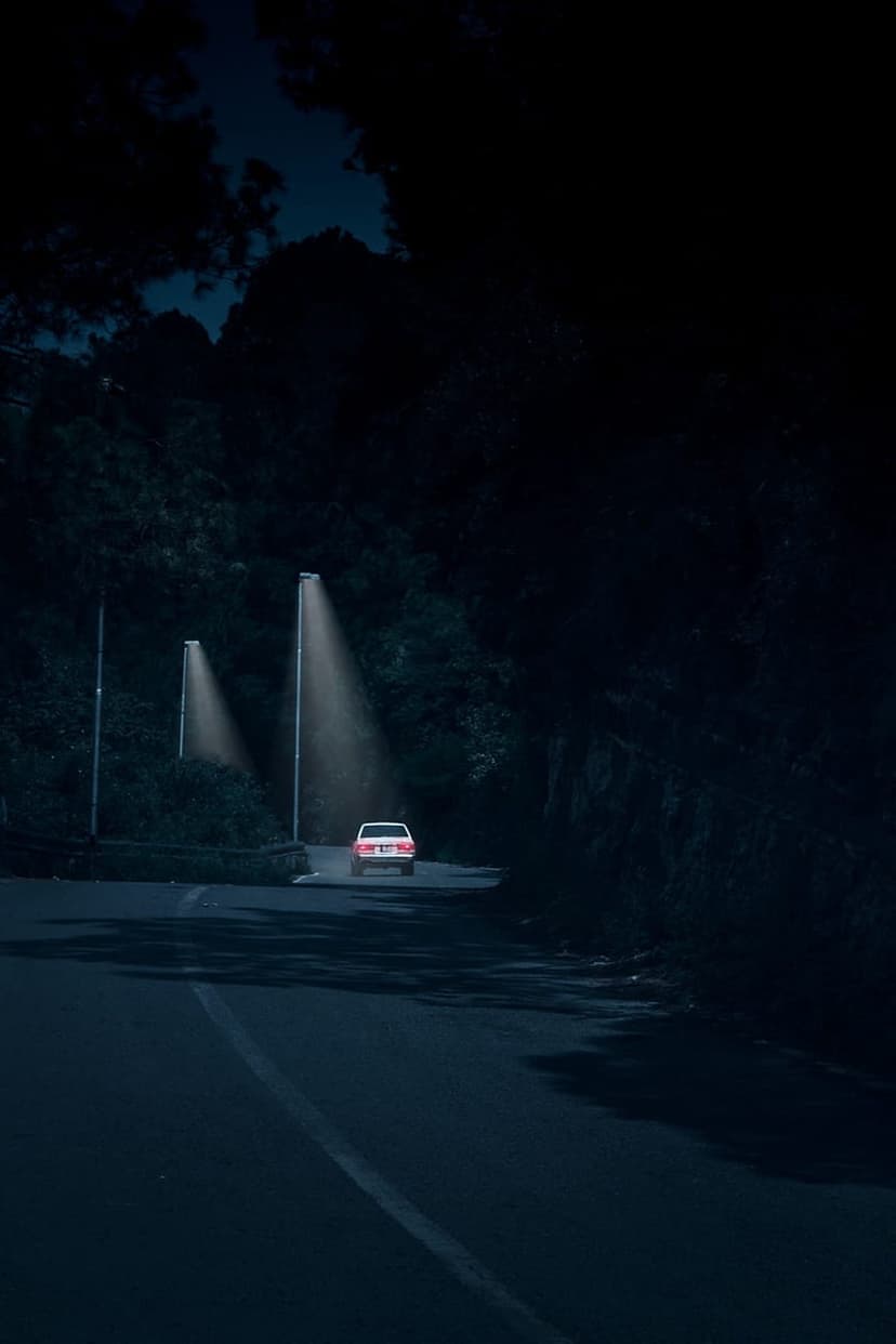 Car with glowing red taillights drives along a winding mountain road flanked by streetlights and dark trees.