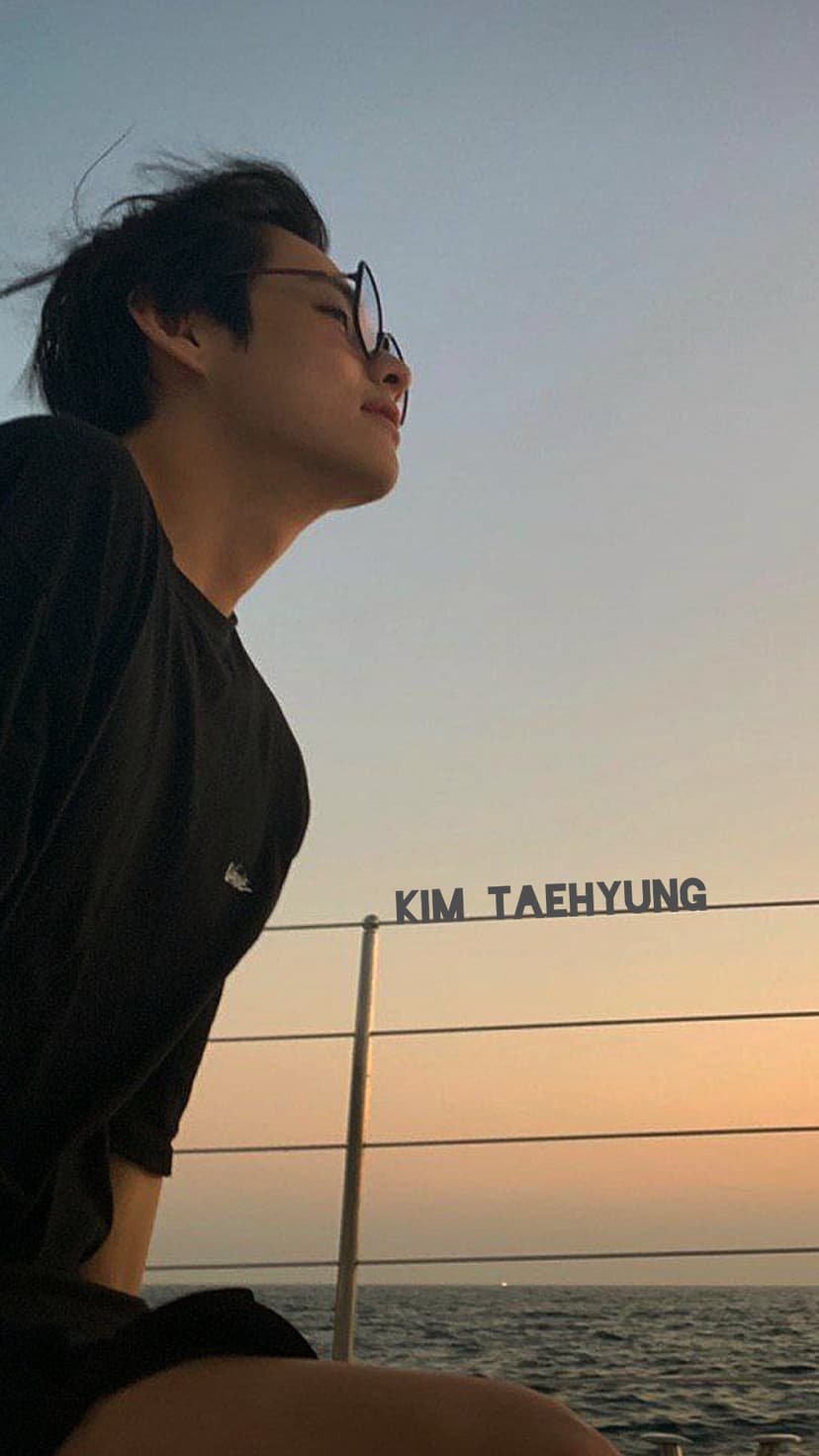 Kim Taehyung wearing glasses looks at a golden sunset over calm ocean waves during a peaceful evening scenery.