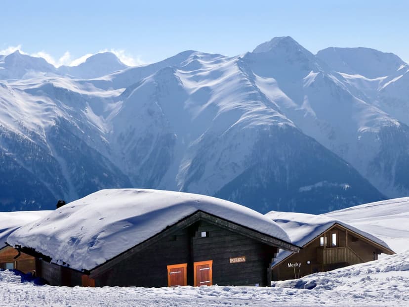 Wooden chalets with glowing windows sit in deep snow before high mountain peaks under a clear blue winter sky.