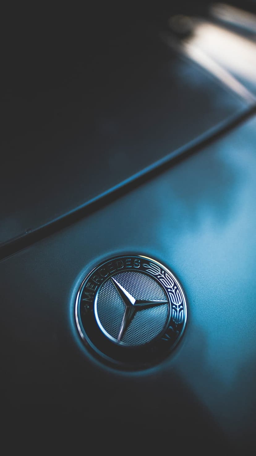 Silver Mercedes-Benz star emblem mounted on a reflective metallic car hood with soft overhead lighting.