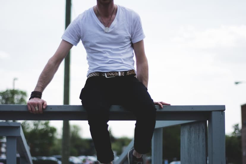 Man wearing a white v-neck and black pants sits on a railing with a soft-focus forest and cloudy sky behind him.