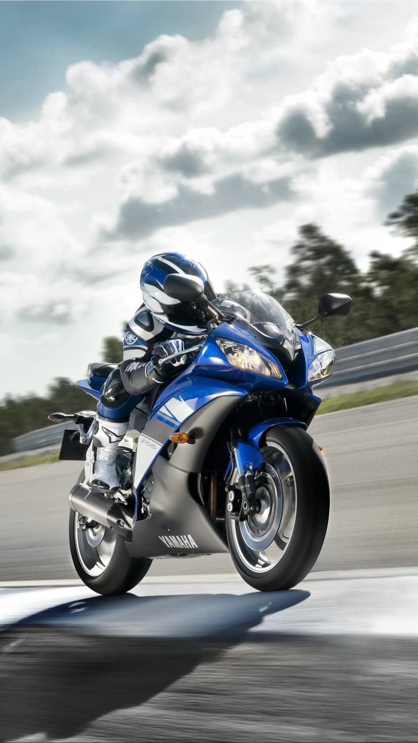 Blue Yamaha R6 motorcycle and rider leaning into a high speed curve on a race track under a cloudy bright sky.