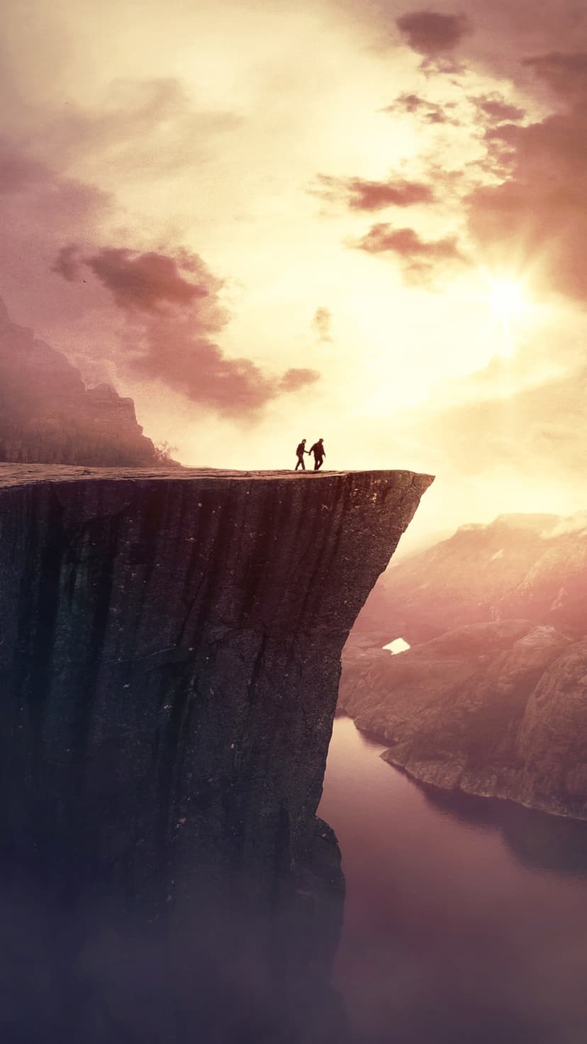 Two silhouetted figures hold hands on a steep cliff overlooking a canyon during a vibrant orange and purple sunset.