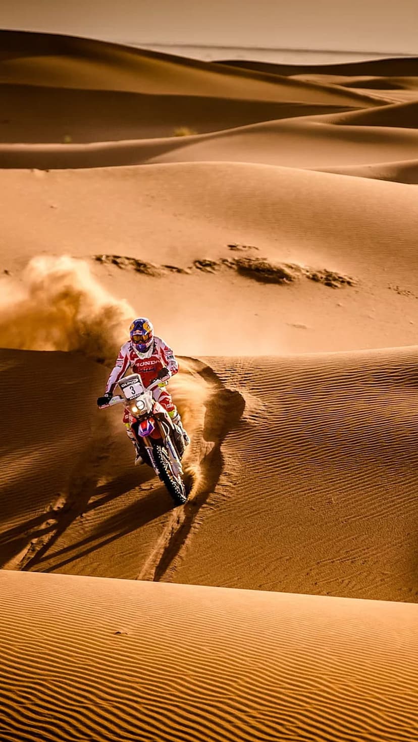 Motorcyclist kicks up a dust plume while racing across vast orange sand dunes under a warm sunset sky.