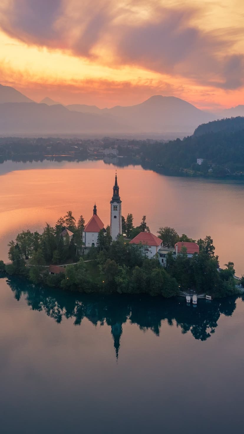 Church of the Assumption of Mary on a small island centered in a still lake under a vibrant sunrise sky.