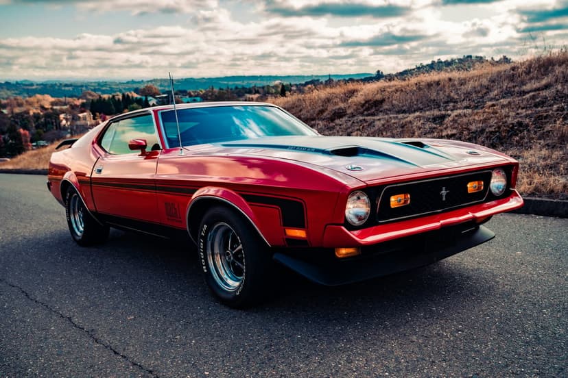 Red 1970s Ford Mustang Mach 1 with black hood stripes parked on a scenic asphalt road under a cloudy sky.