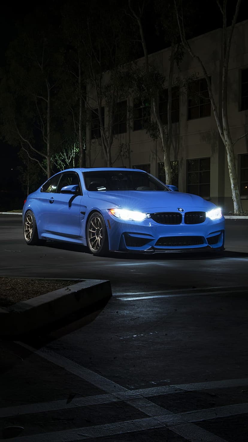 Blue BMW M4 coupe parked on dark asphalt at night with glowing white headlights and a building in the distance.