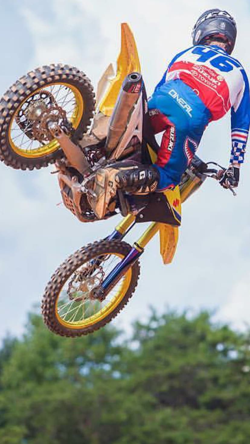 Motocross rider wearing blue and red gear jumps a yellow dirt bike against a cloudy sky with blurred trees.