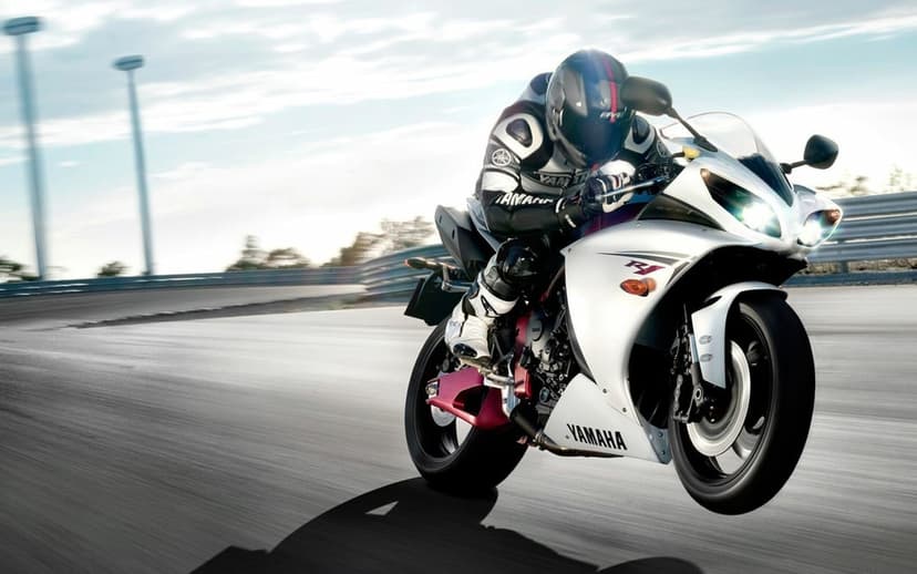 White Yamaha R1 sportbike performing a wheelie on a track with a rider in full gear and heavy motion blur.