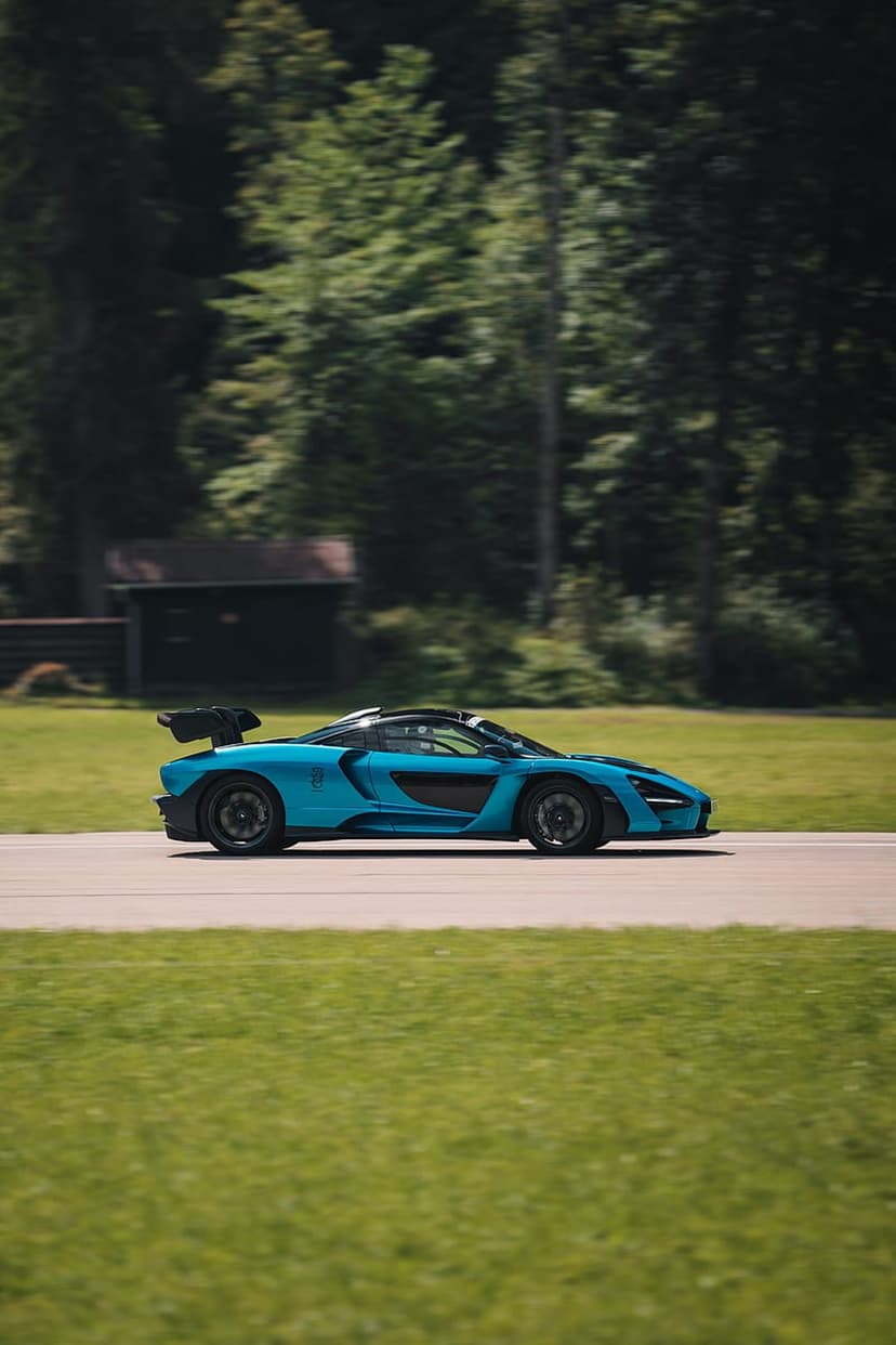 Blue McLaren Senna hypercar speeds on a sunlit race track with green trees and grass in the background.