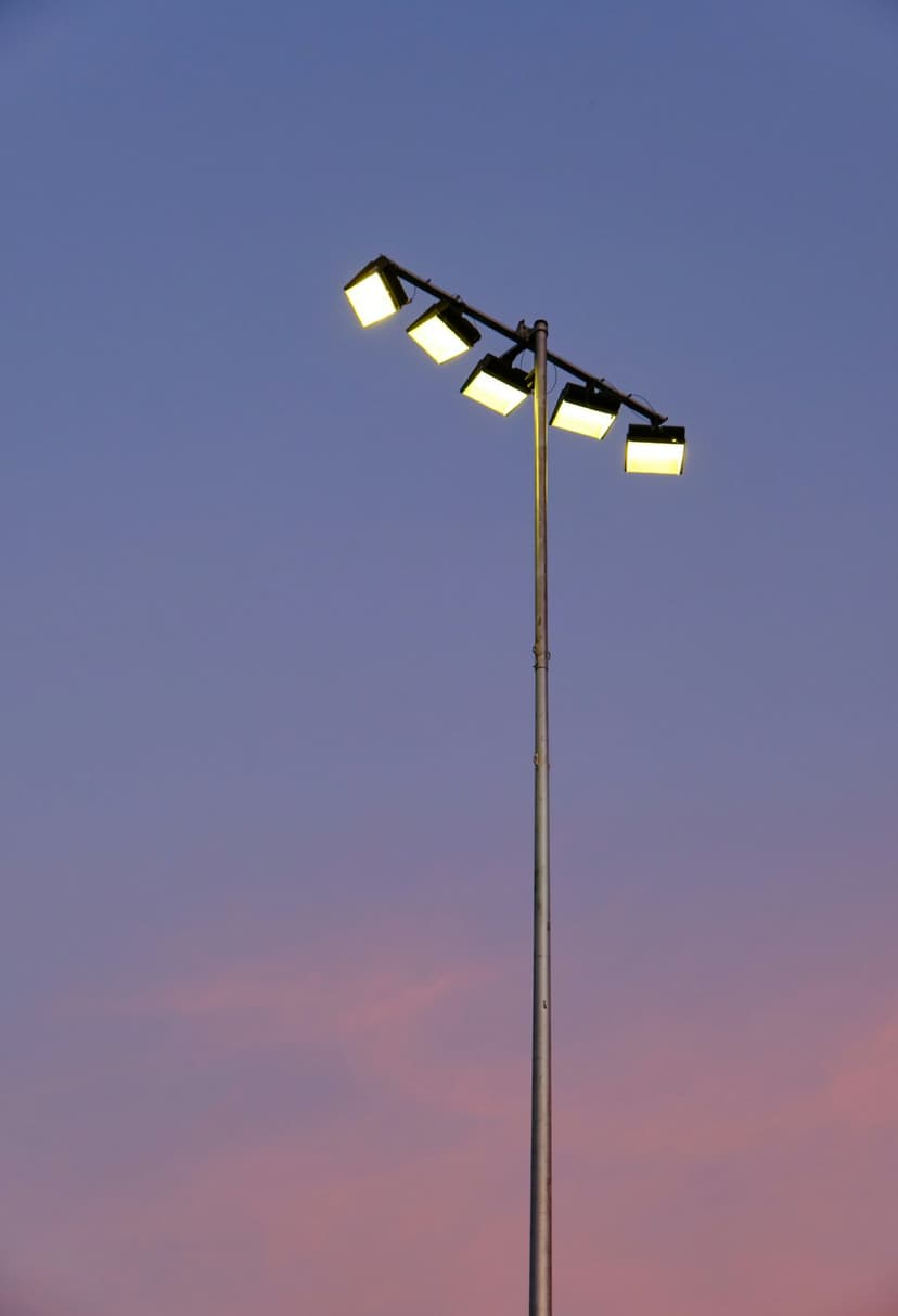 Metallic floodlight pole with glowing yellow lamps against a gradient sky of dark blue, purple, and soft pink.