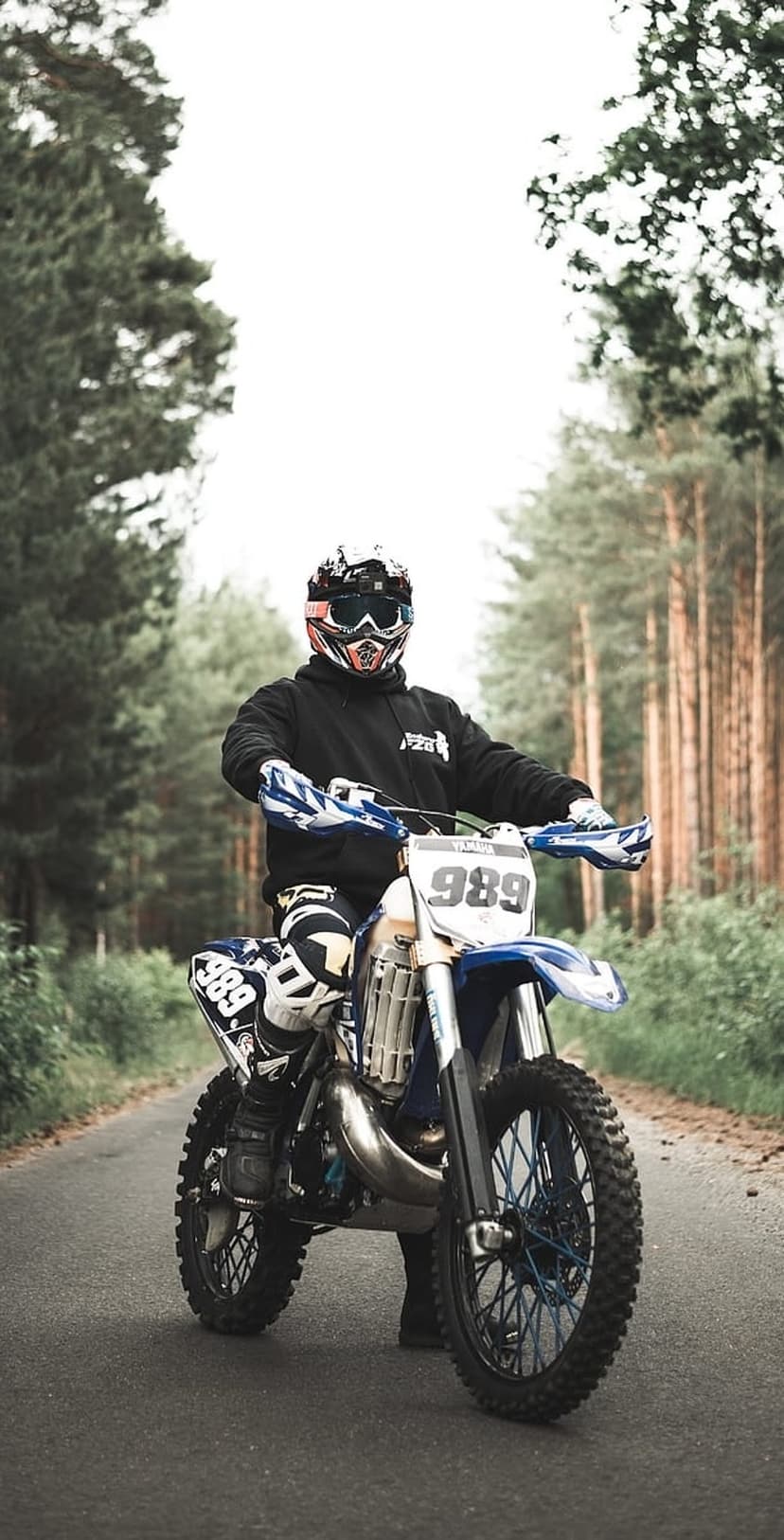 Motocross rider on a blue Yamaha dirt bike numbered 989 parked on a paved road surrounded by dense pine trees.