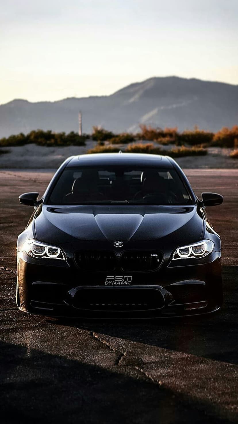 Black BMW M5 with glowing headlights parked on a sandy desert road under a hazy, dusky sky during twilight.