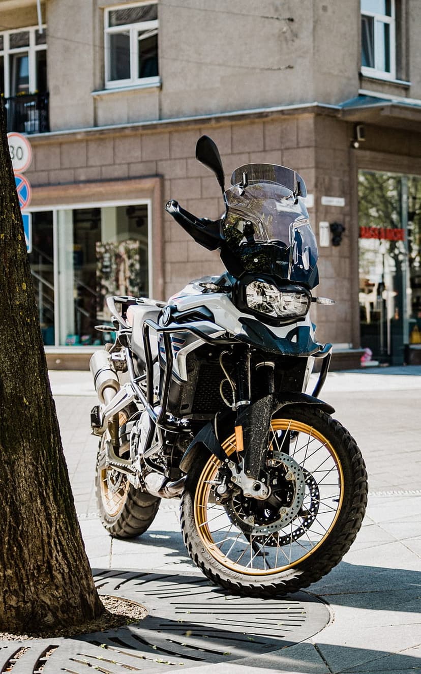 BMW adventure motorcycle with gold rims parked on a city sidewalk next to a tree trunk and urban storefront.