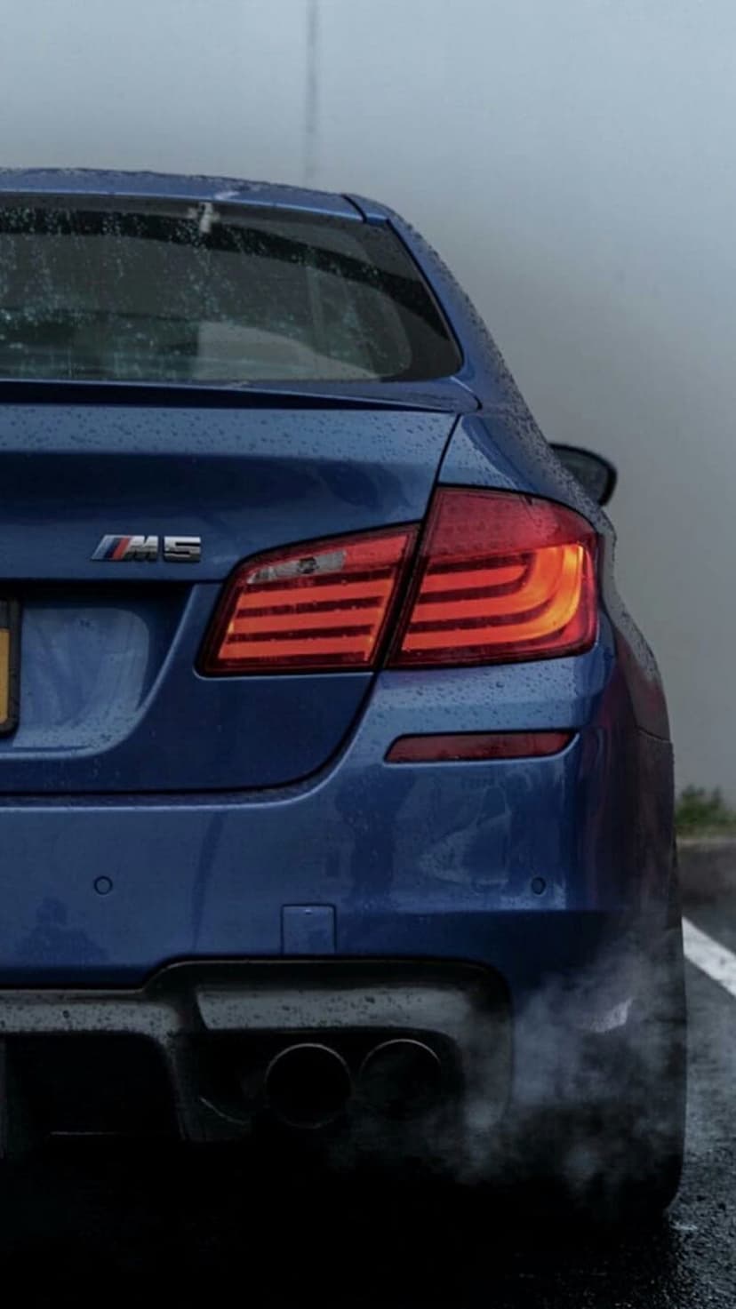 BMW M5 rear bumper and quad exhaust pipes releasing white fumes on a wet asphalt road during a rainstorm.