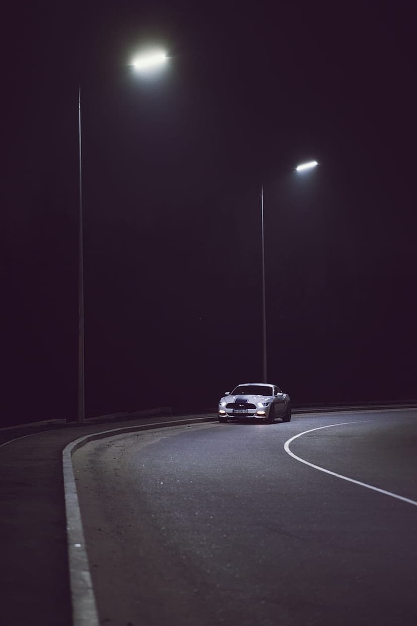 White Ford Mustang sports car cruising a dark curved road at night illuminated by overhead street lamps.