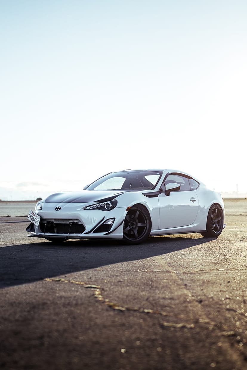 Toyota GT86 with a white paint job and black hood accents parked on dark asphalt under a bright clear sky.