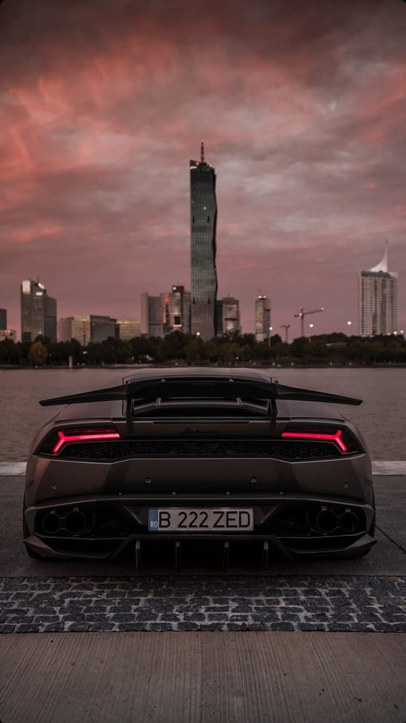 Black Lamborghini rear view with red taillights parked on cobblestone before a sunset city skyline silhouette.