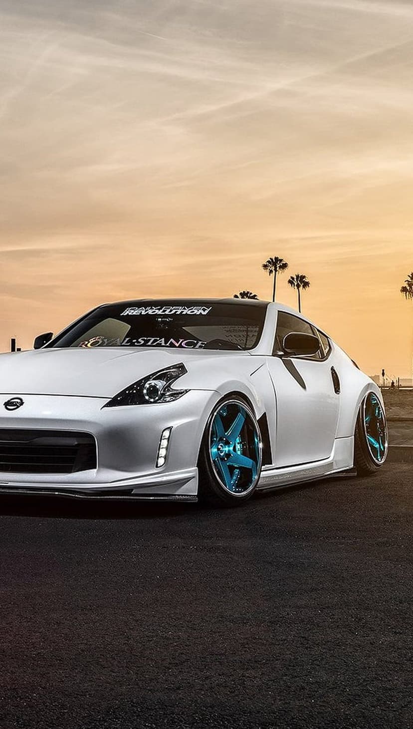 White Nissan 370Z with teal wheels parked near silhouetted palm trees under a vivid golden hour sunset sky.