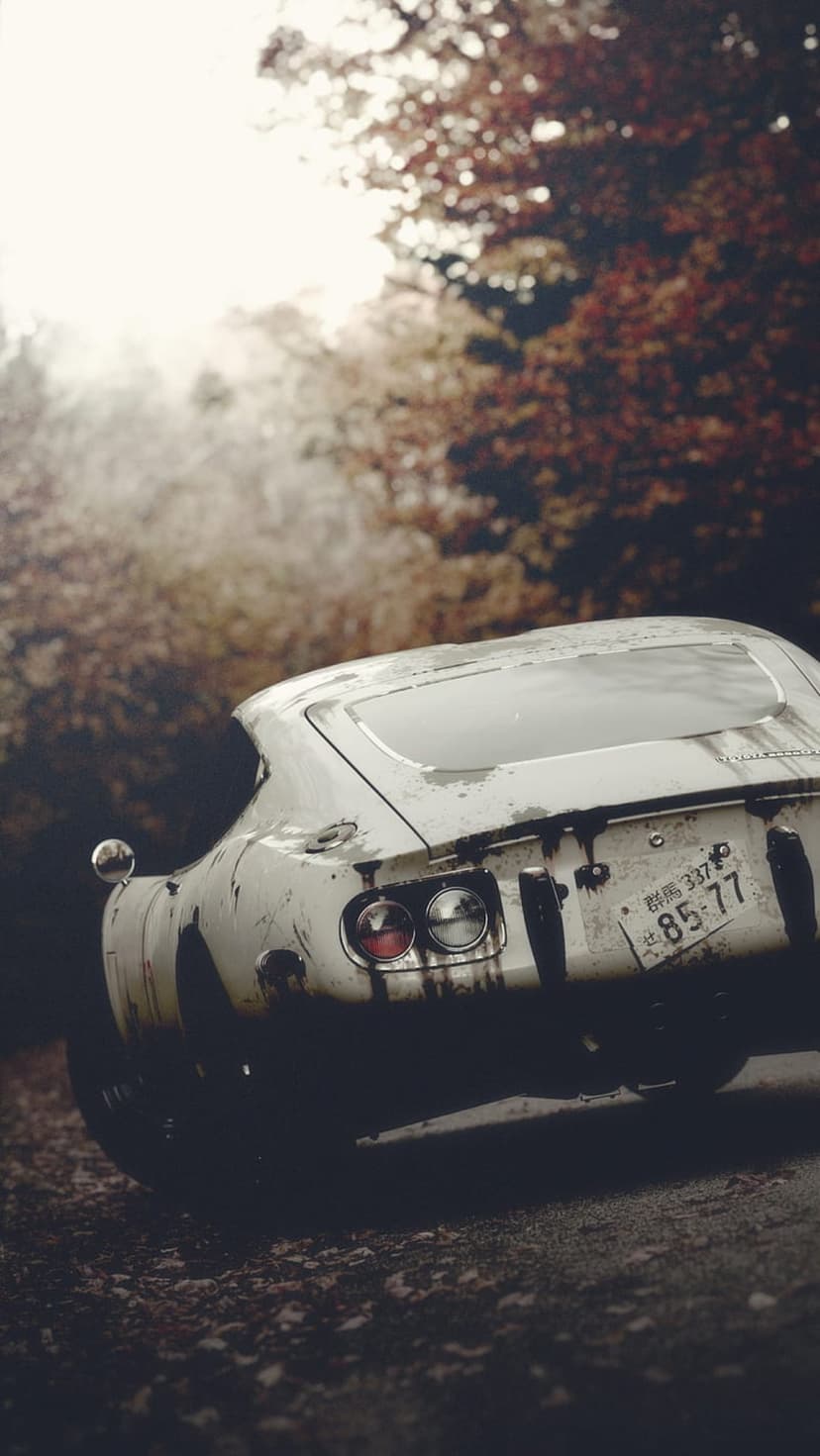 Rusted white vintage car parked on a dirt road covered in orange autumn leaves within a sunlit forest.