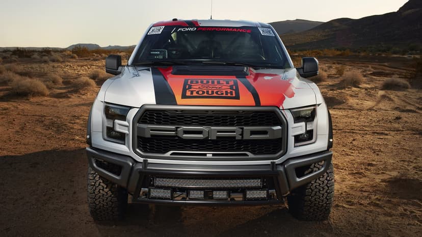 Front view of a white and red Ford Raptor racing truck with a black grille and light bars in a desert setting.