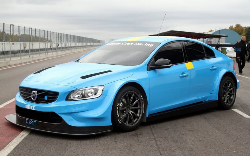 Cyan blue Volvo Polestar racing car with a wide body kit parked on an asphalt racetrack under a grey sky.