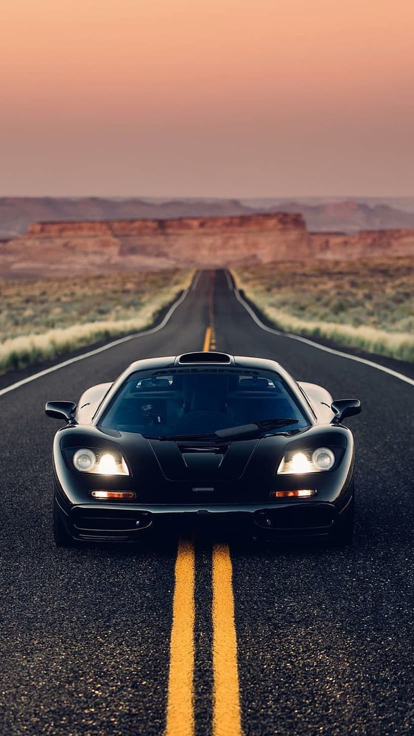 Black McLaren F1 supercar with lit headlights driving on a desert road under a vibrant sunset sky.