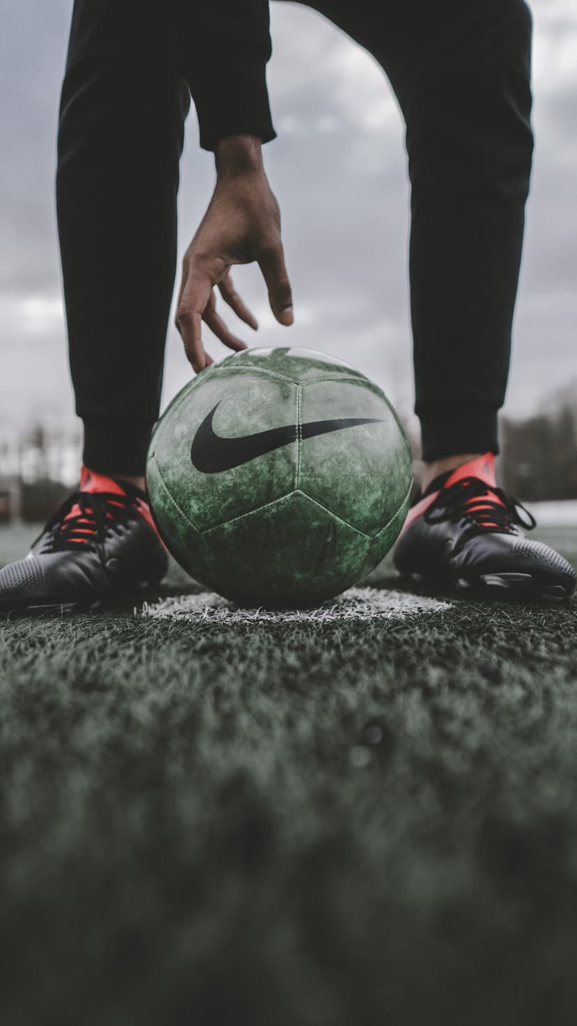 Soccer player's hand reaching toward a green Nike ball on a grassy pitch with red-accented cleats visible.