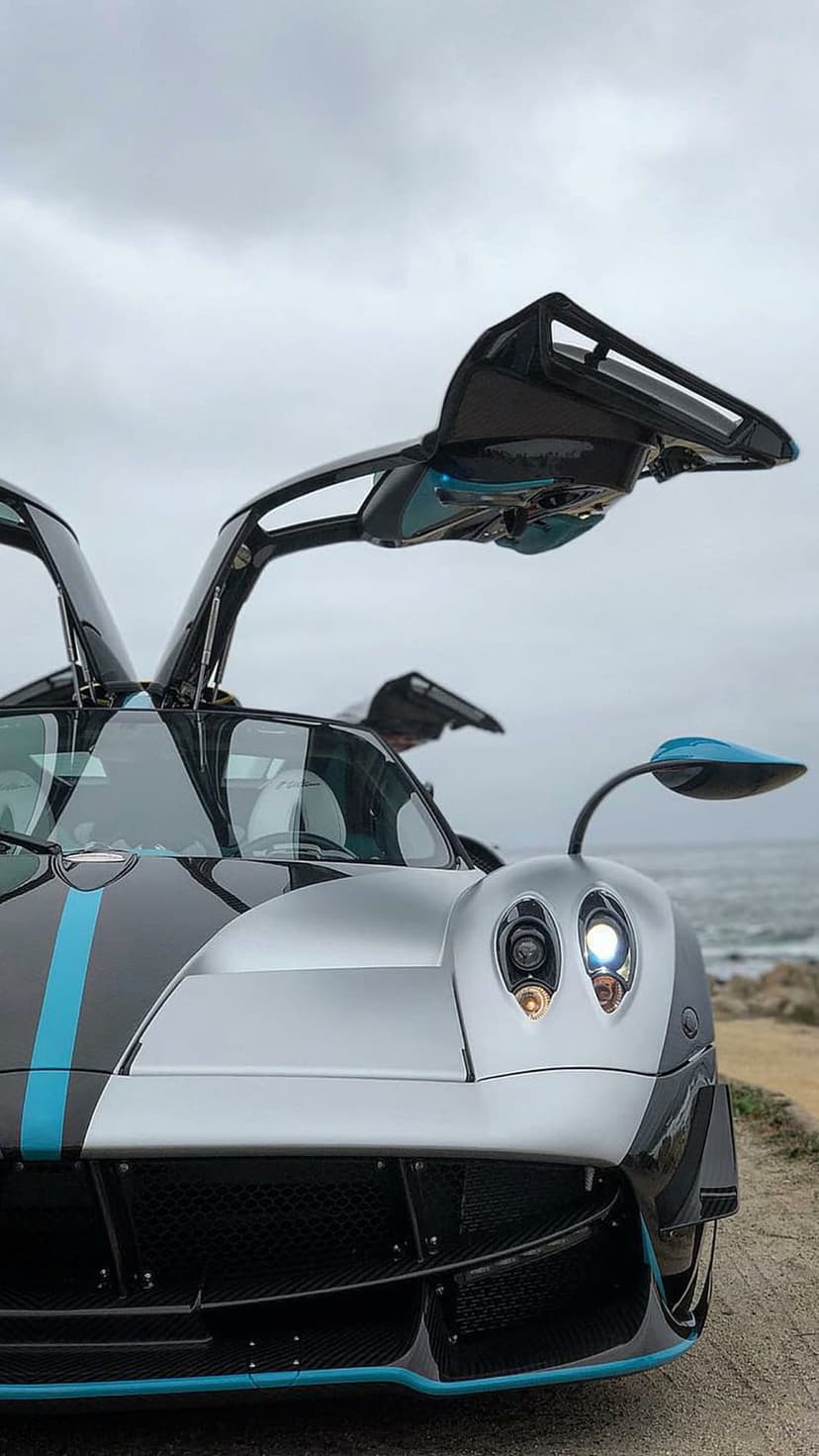 Pagani Huayra Roadster with open doors on a cliffside road under a cloudy sky during a luxury coastal drive.