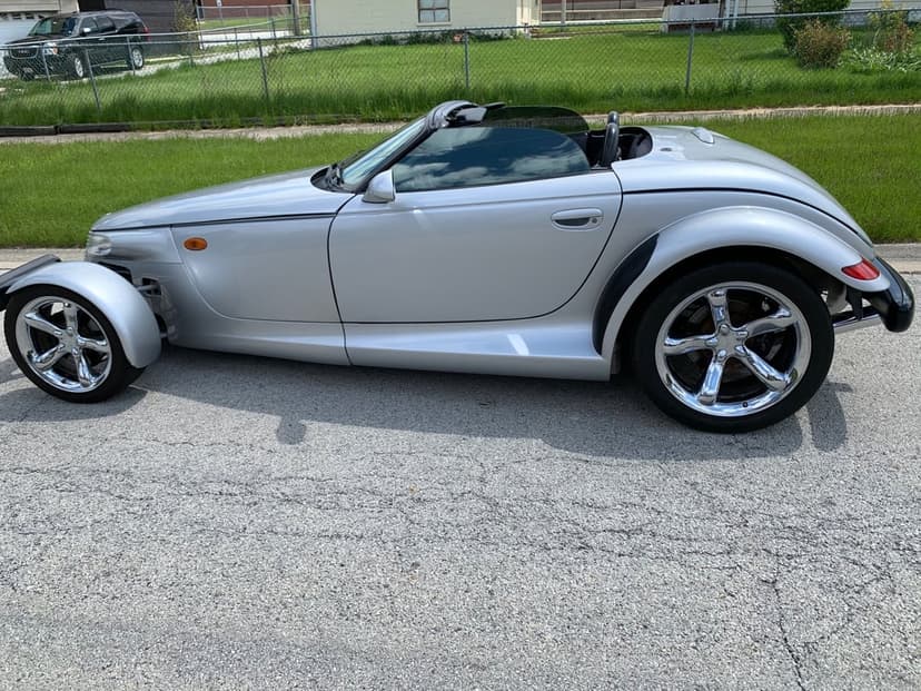 Side view of a silver Plymouth Prowler roadster with chrome wheels parked on an asphalt street in daylight.
