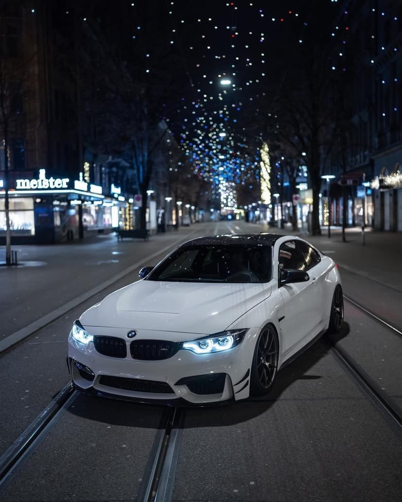 White BMW M4 coupe driving under festive string lights on a city street at night with glowing headlights.