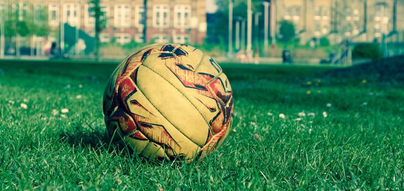 Vintage soccer ball on vibrant green grass with a blurred city skyline and trees in a warm afternoon light.