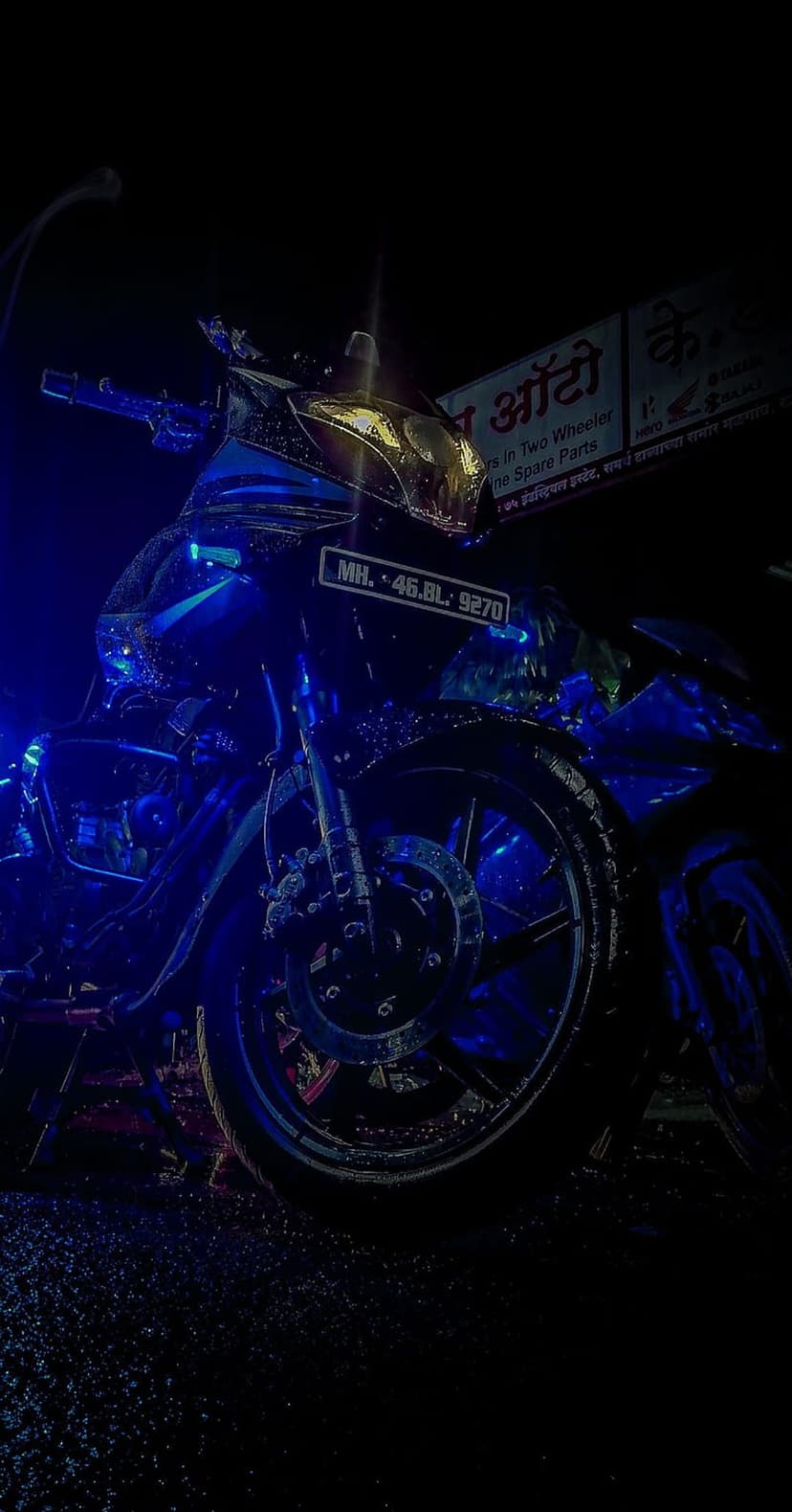 Black motorcycle with blue neon underglow and yellow headlights sits on a wet, reflective street at night.