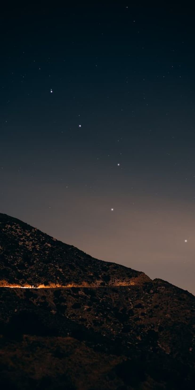 Winding mountain road glowing under a starlit indigo sky with jagged peaks silhouetted against a soft horizon glow.