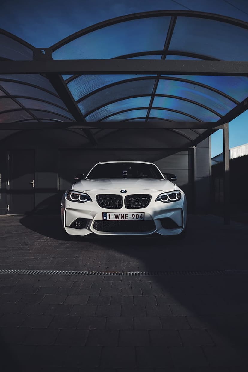White BMW M2 parked under a modern arched carport with a blue twilight sky visible through translucent roof panels.