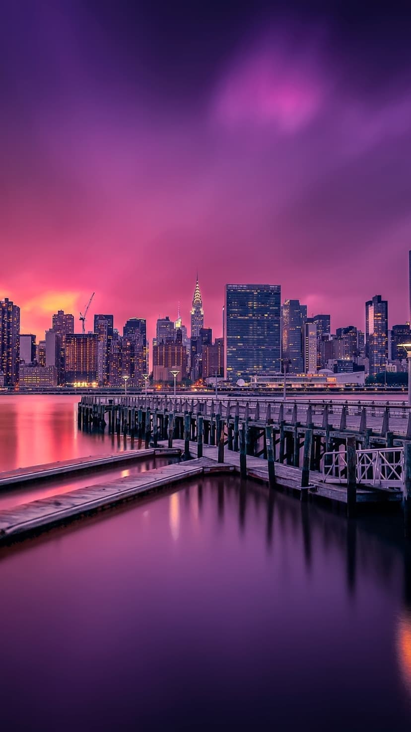 Manhattan skyline lights reflect in calm harbor water behind weathered wooden piers during a purple twilight.