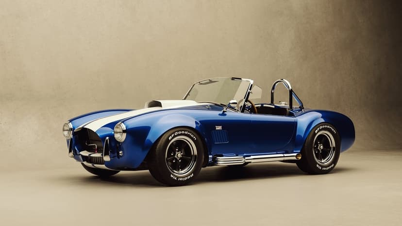 Blue Shelby Cobra sports car with white racing stripes and chrome side pipes against a beige studio background.