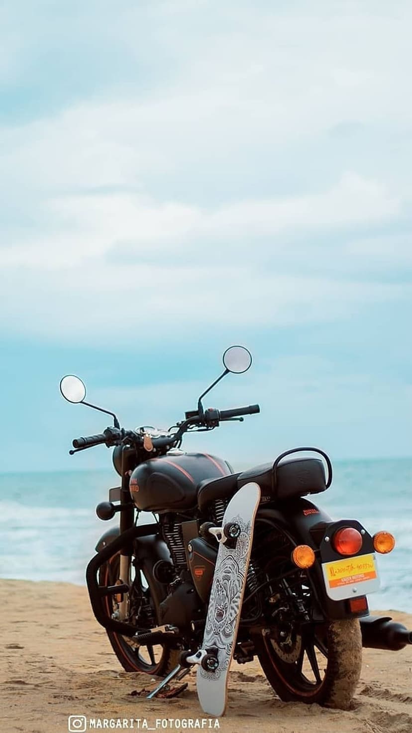 Black motorcycle and white skateboard lean together on a sandy beach with ocean waves under a pale blue sky.
