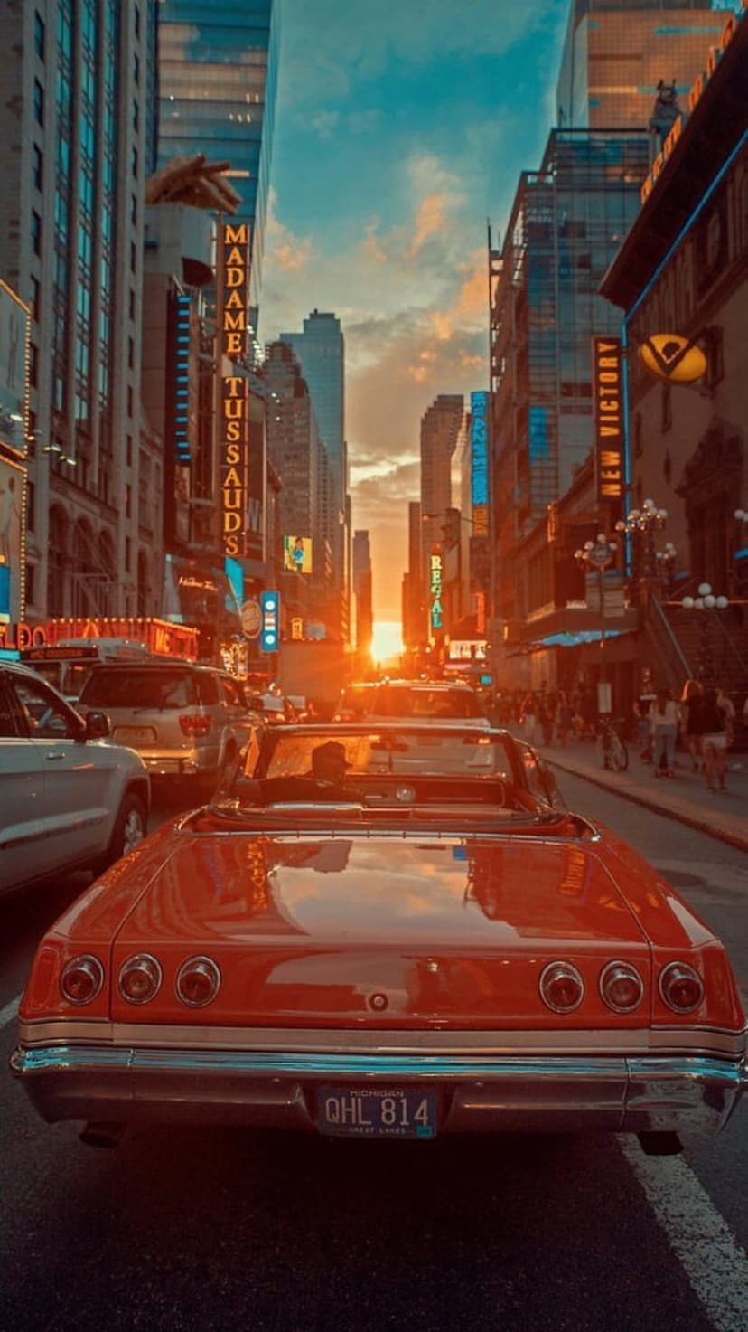 Red vintage convertible drives through Times Square at sunset with bright neon theater signs and golden light.