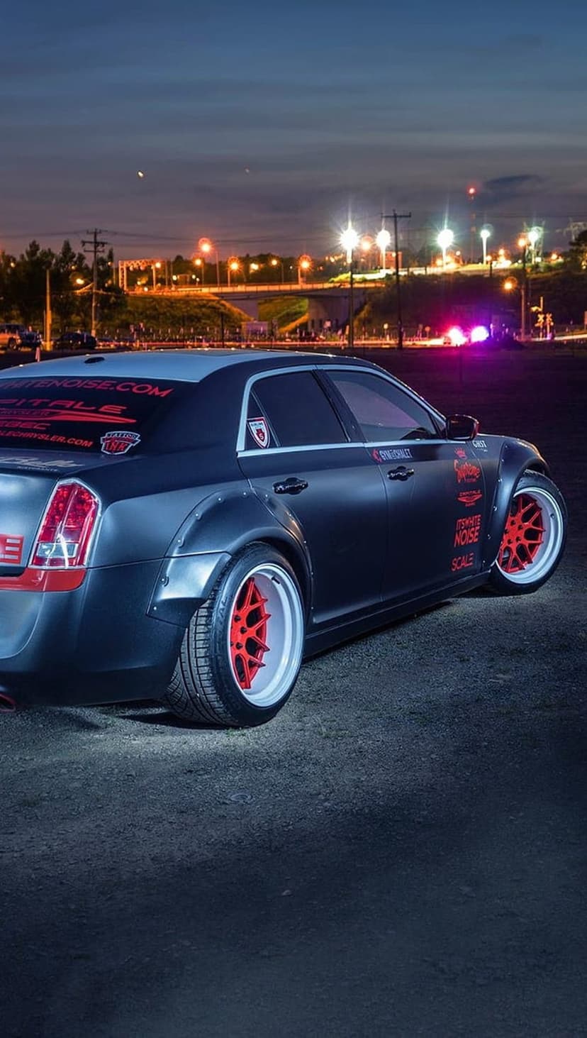 Matte black Chrysler 300C with widebody kit and red rims parked on dark asphalt under city bridge lights at night.